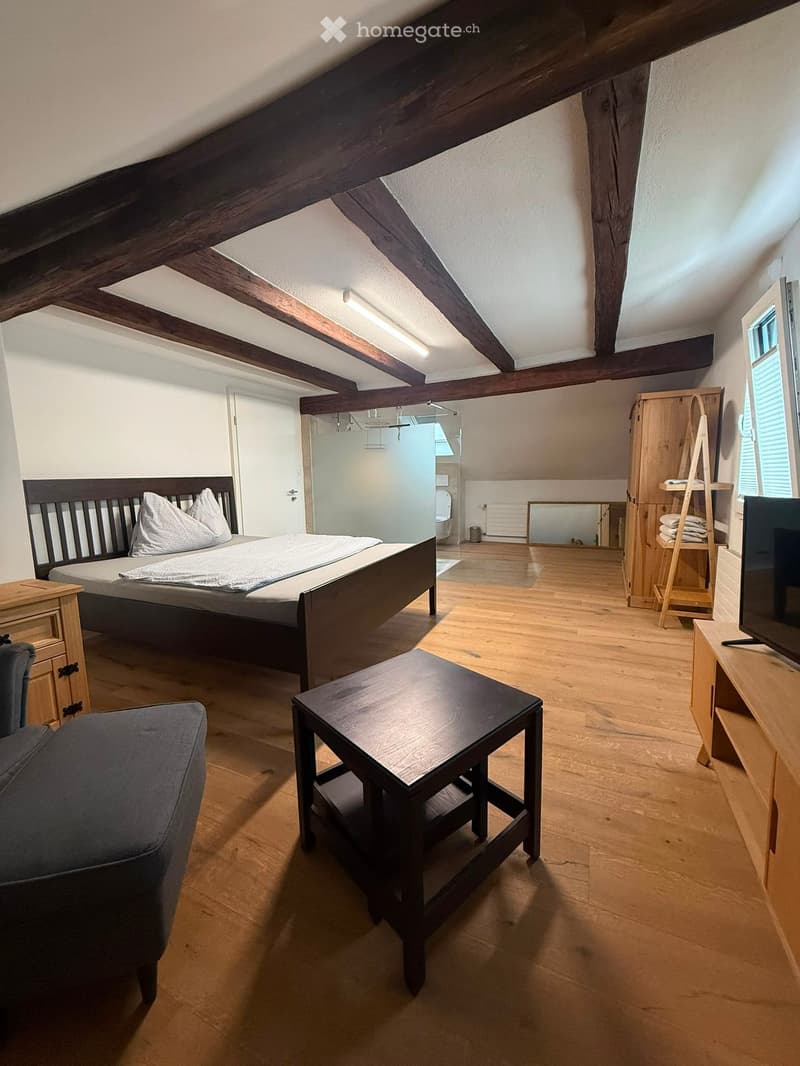 Cozy attic bedroom with wooden beams, a double bed, nightstand, sofa, small table, TV, and a wooden wardrobe.
