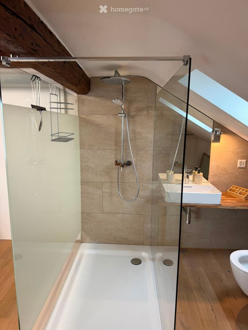 Modern bathroom with a large bathtub, glass-enclosed shower, and a sleek sink area. Features wooden flooring and exposed wooden beams on the ceiling.