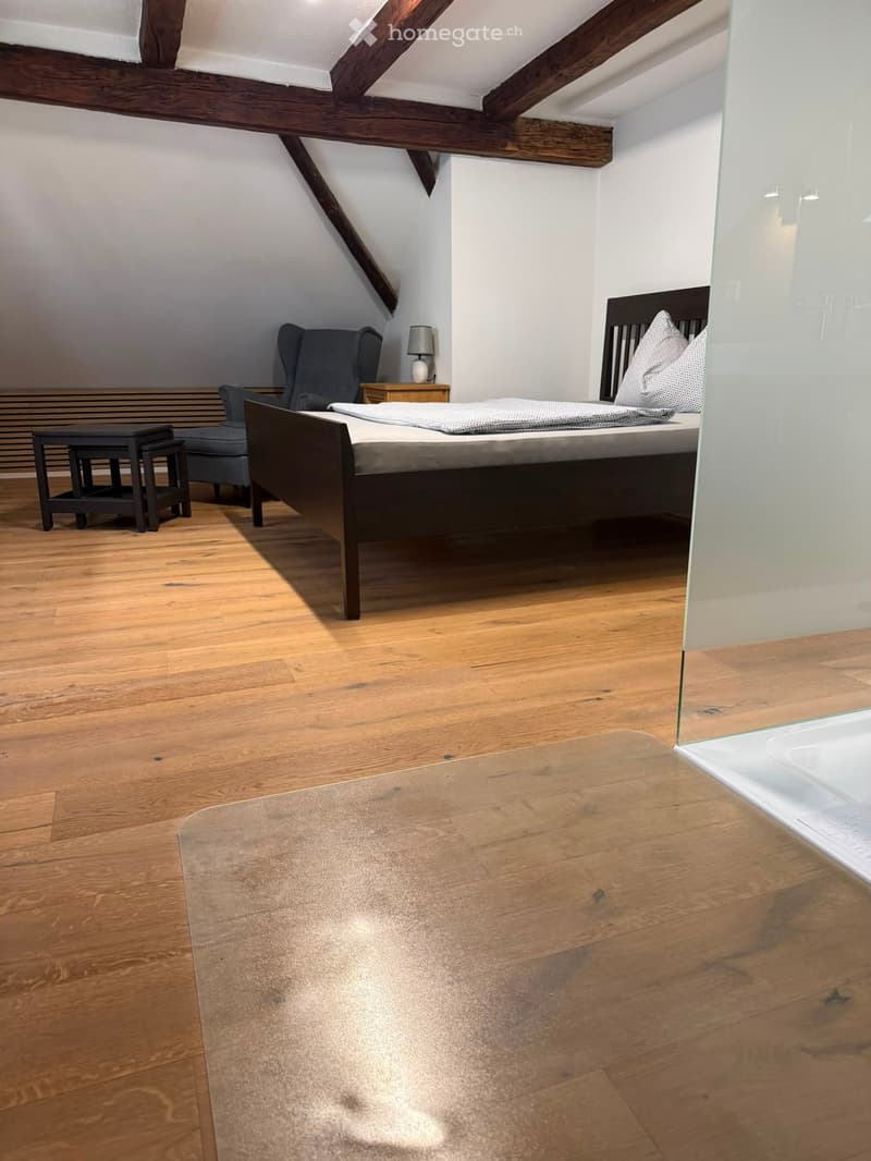 Cozy attic bedroom with wooden floors, slanted wooden ceiling beams, a double bed with pillows and a blanket, a nightstand with a lamp, an armchair, and a small table.