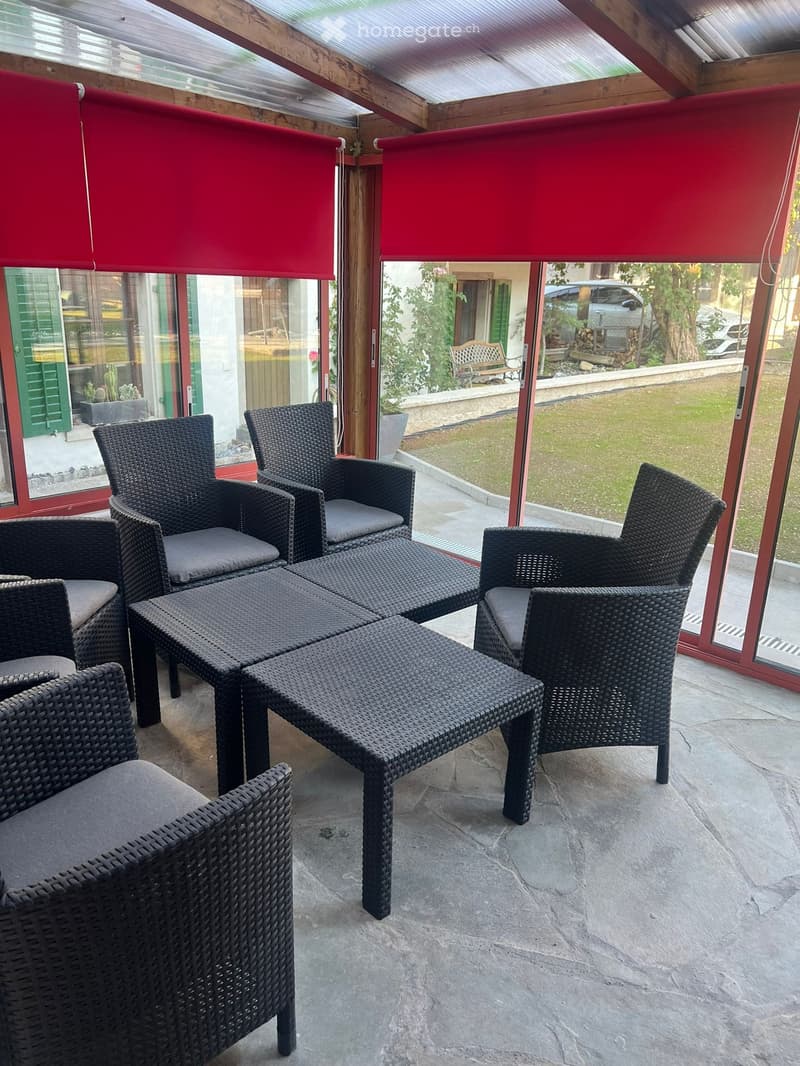 A covered outdoor area with wicker furniture, including a table and chairs, large windows with red blinds, and a view of a garden.