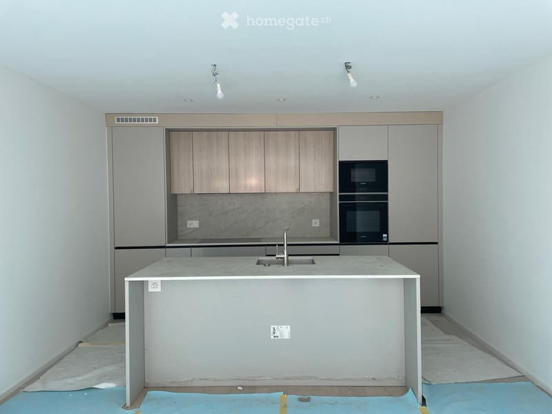 Modern kitchen with light wood cabinets, a central island, and built-in appliances including an oven and microwave. The countertops are light-colored, and the floor is covered with protective sheeting.