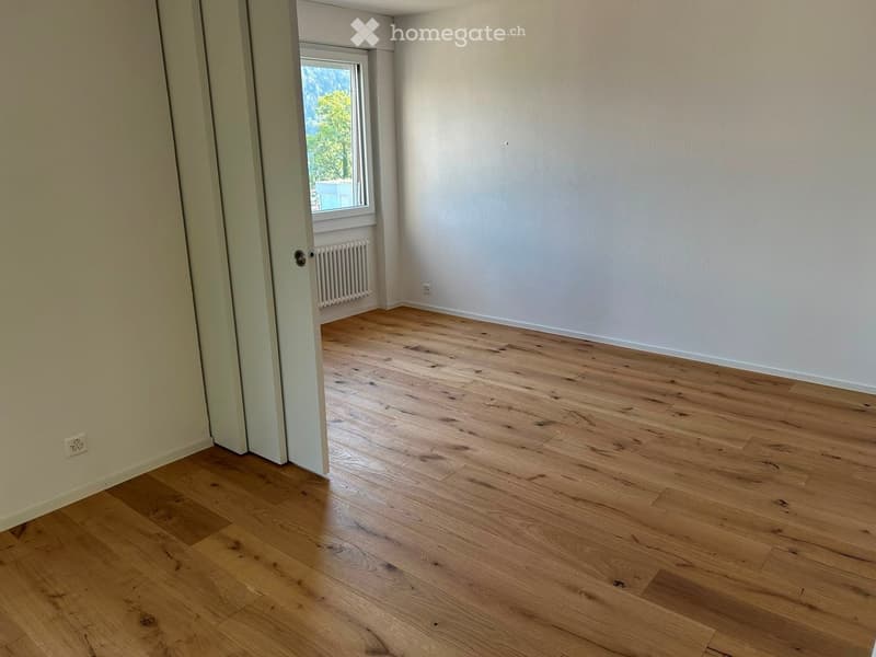 Empty room with light wooden flooring, white walls, and a window showing greenery outside. No furniture or equipment is present.