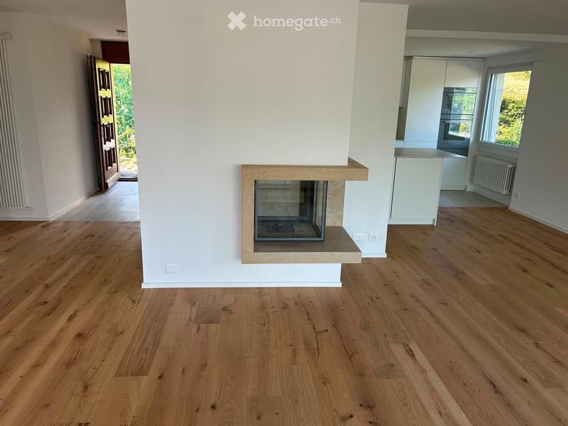 Modern living room with light wooden flooring, a built-in fireplace, and a view of a kitchen area with white cabinets and a window.