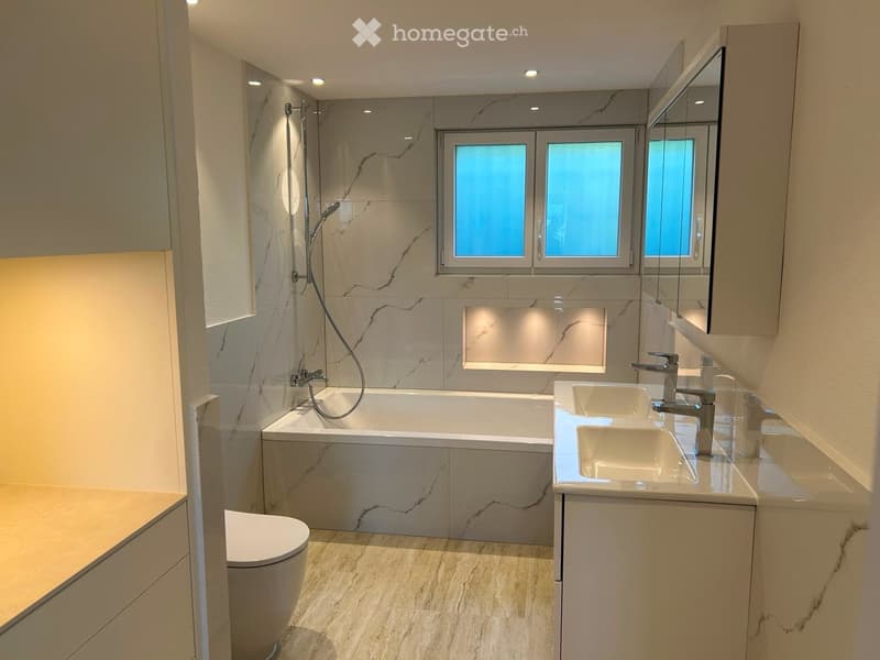 Modern bathroom with marble tiles, a bathtub, toilet, double sink, and a large mirror. Features recessed lighting and a window with frosted glass.