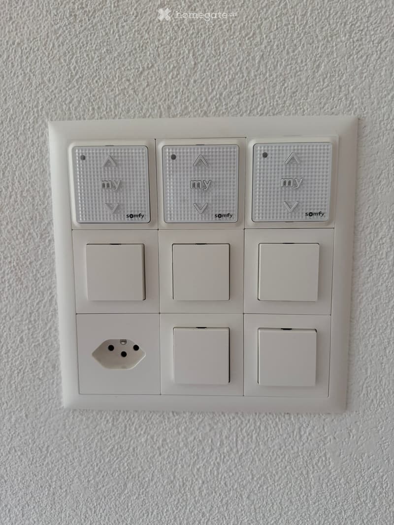 A white wall-mounted smart home control panel with multiple touch-sensitive buttons and a power outlet.