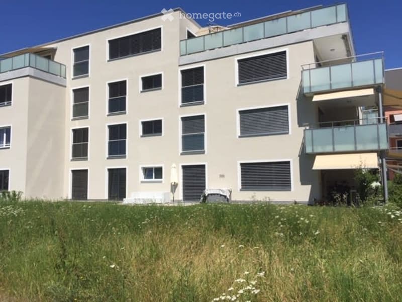 Modern multi-story residential building with balconies and large windows, surrounded by a grassy area with small white flowers.