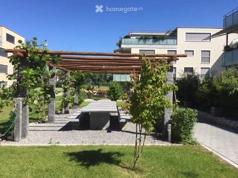 Well-maintained garden with a wooden pergola, seating area, and lush greenery, surrounded by modern buildings.