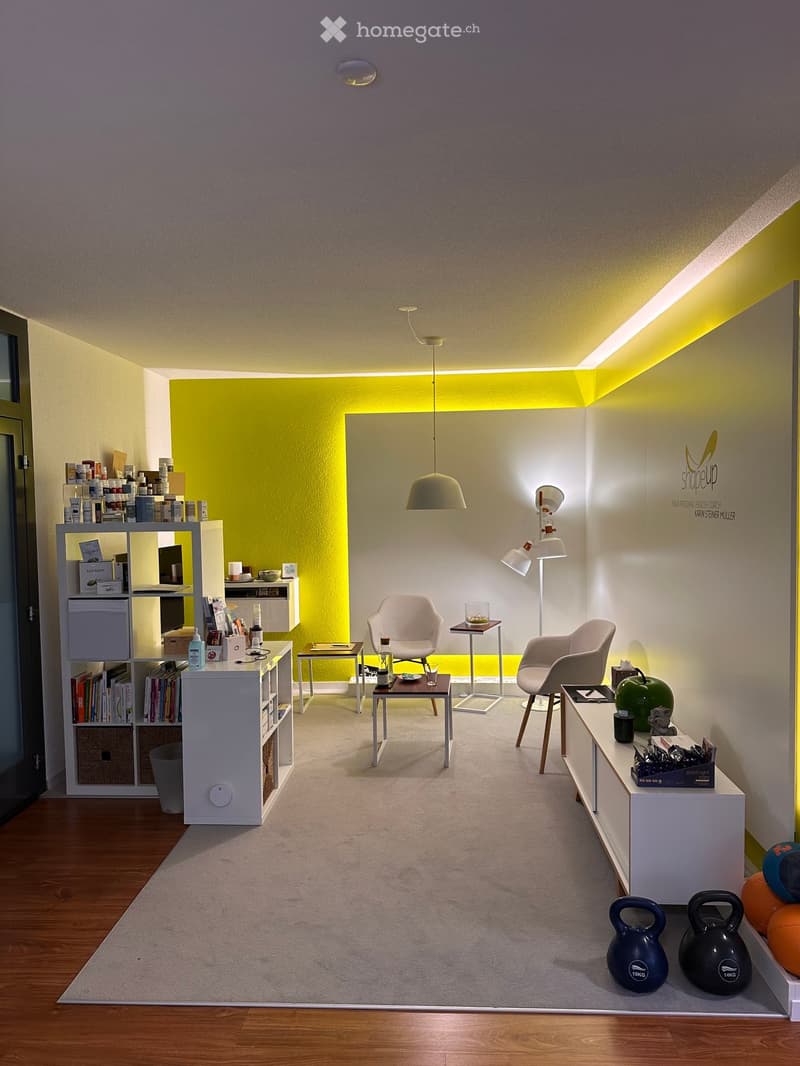 Modern, well-lit room with a yellow accent wall, white furniture, and exercise equipment including kettlebells and a ball. Features a shelf with books and products, a small table with a lamp, and a white chair.