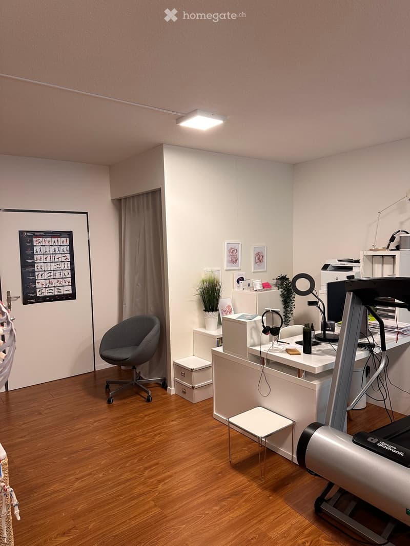Modern office space with wooden flooring, white desk, grey chair, treadmill, and various office equipment.