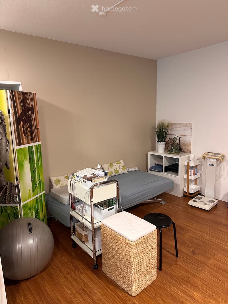 A well-lit room with a bed, a wicker stool, a large exercise ball, a rolling cart with supplies, a small table with a plant and decorative items, and a black chair.
