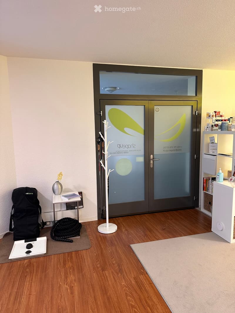 A well-lit entryway with wooden flooring, a coat rack, a small table with a vase and books, a backpack, a rope, and shelves with various items.