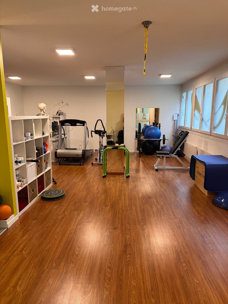 Well-lit fitness room with wooden flooring, various exercise equipment including a treadmill, elliptical machine, exercise balls, a mirror, and a shelf with fitness accessories.