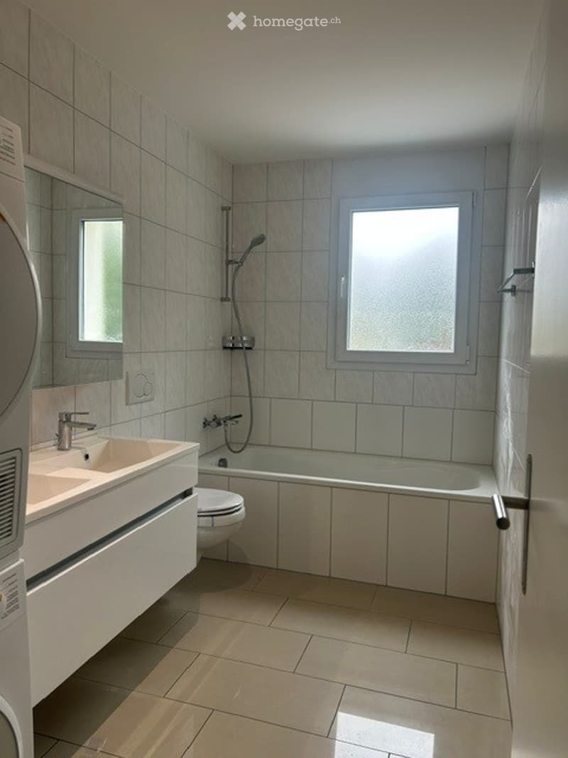 Modern bathroom with a white tiled wall, a large window, a sink with a faucet, a mirror, a toilet, and a bathtub with a showerhead.