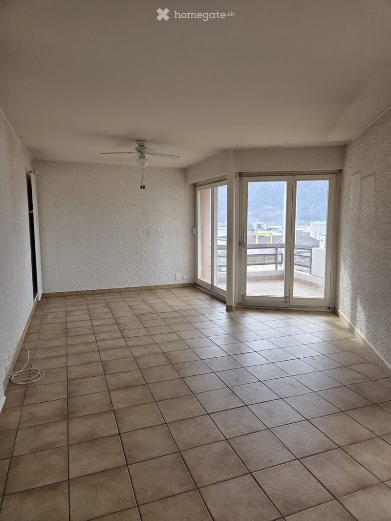 Empty room with white walls, brown tiles, a ceiling fan, sliding doors, a balcony with a fence