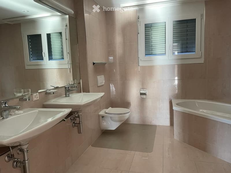 Modern bathroom with two sinks, a toilet, a bathtub, and a large mirror. Light-colored tiles and a window with blinds are visible.