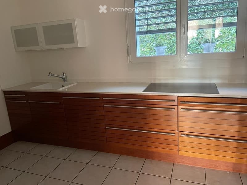 Modern kitchen with wooden cabinets, a sink, a faucet, and a microwave above the counter. The space features tiled flooring and a large window with blinds.