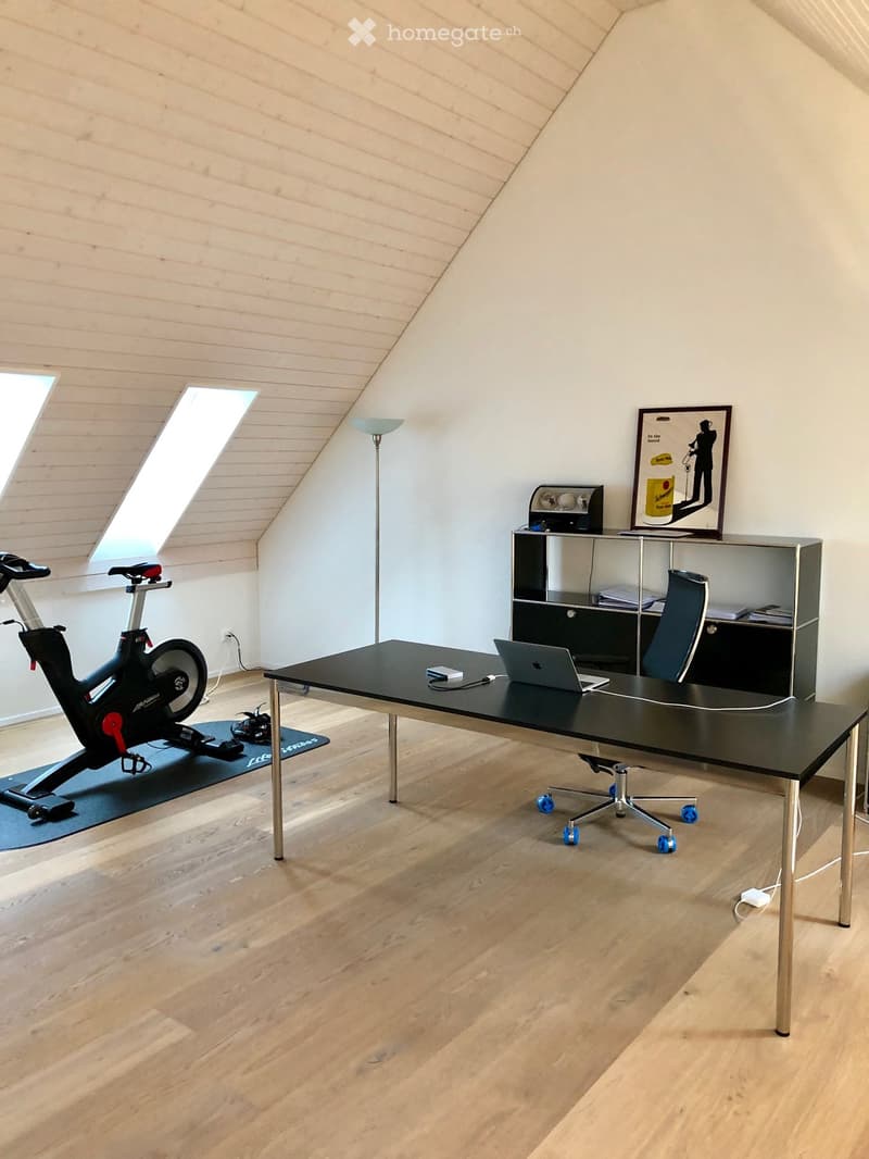 Modern, well-lit room with wooden flooring, a sleek black desk, a laptop, a rolling chair, an exercise bike, a cabinet, and a floor lamp.