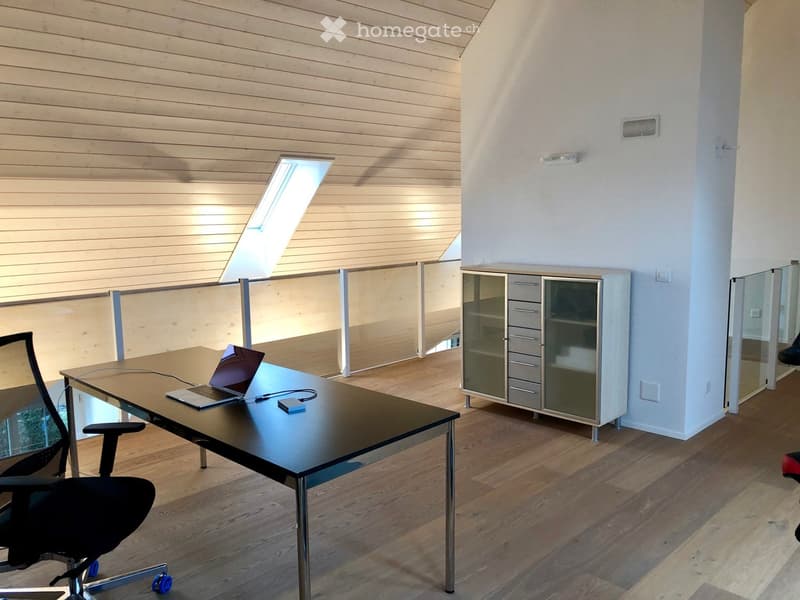 Modern office space with a sleek black desk, ergonomic chair, laptop, and a white cabinet with glass doors. The room features wooden flooring, white walls, and a large skylight providing natural light.