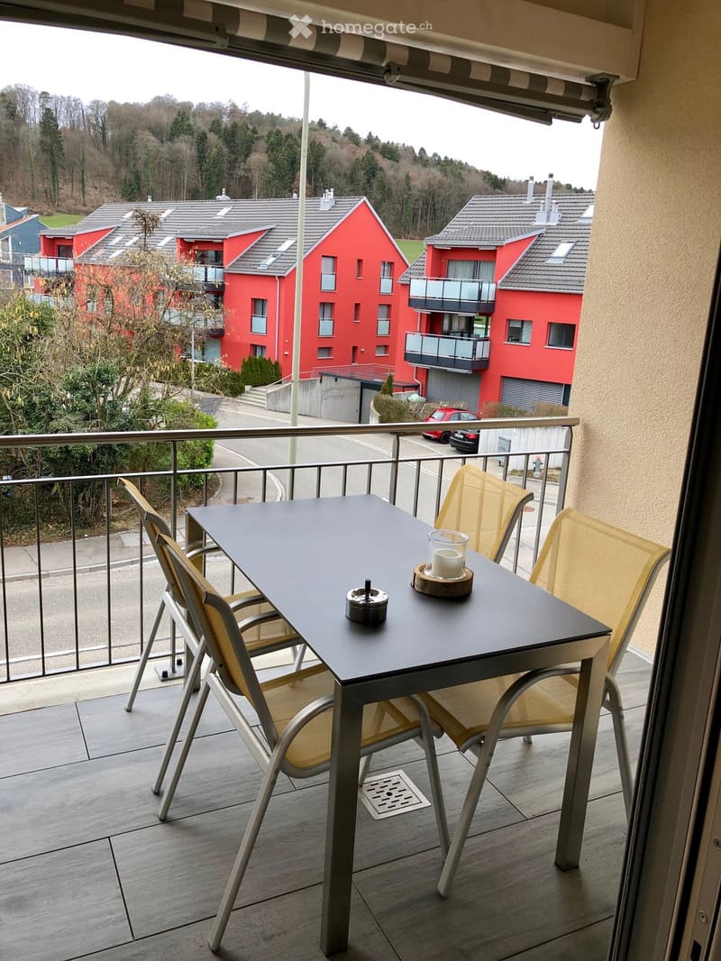 Balcony with a small table and two chairs, overlooking a residential area with red buildings and trees.