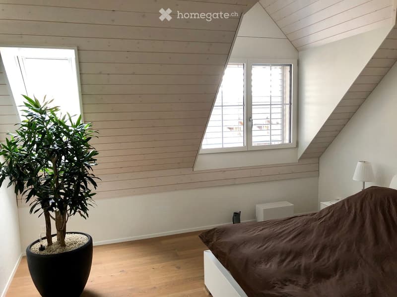 Cozy bedroom with wooden floors, slanted ceiling, large windows, a bed with brown bedding, a table lamp, and a potted plant in the corner.