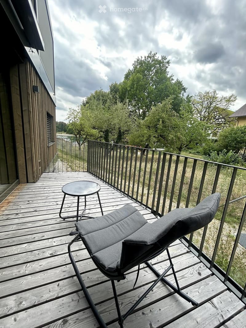 Wooden deck with a lounge chair, small round table, and metal railing overlooking a green landscape.