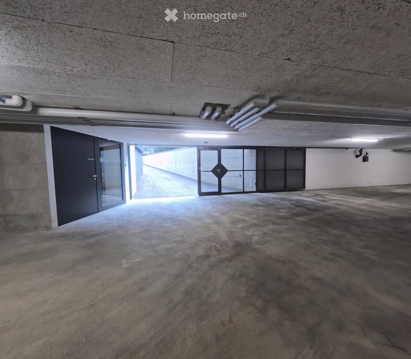 Empty underground parking space with concrete floors and walls, featuring a large open area, a few doors, and some lighting fixtures.