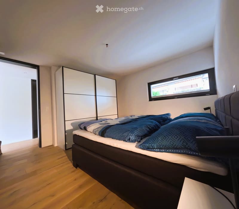 Modern bedroom with a large bed, dark blue bedding, and a white wardrobe. The room features wooden flooring and a window with a view.