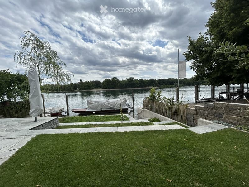 A serene garden area with a well-maintained lawn, stone pathways, and a view of a calm river with docked boats. The area includes a wooden fence, a covered patio umbrella, and a stone staircase leading to the water.