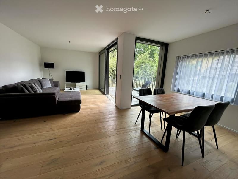 Open-plan living and dining area with wooden flooring, a grey sectional sofa, a coffee table, a TV on a stand, a dining table with chairs, and large windows with sheer curtains leading to a balcony.
