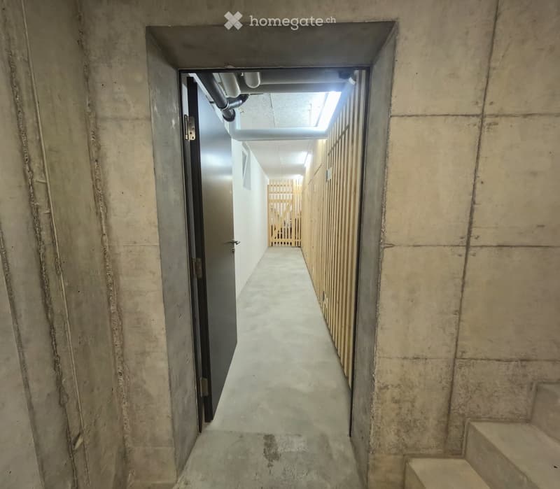 A narrow concrete corridor with a door on the left and a wooden barrier on the right, leading to a brighter area with visible pipes and a small window at the end.