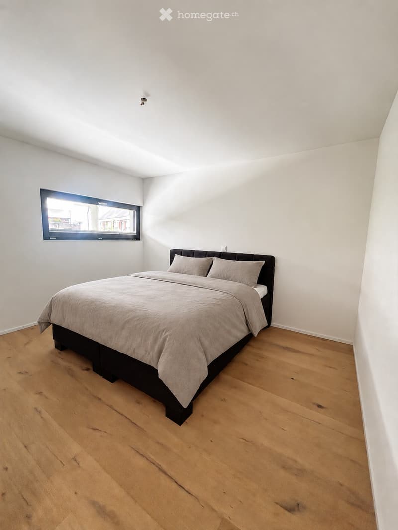 A modern bedroom with a wooden floor, a large window, and a neatly made bed with pillows and a duvet.