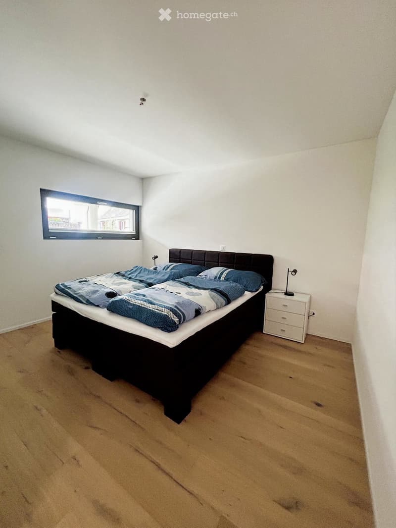 Bedroom with wooden flooring, a large window, a double bed with blue bedding, a nightstand with a lamp, and a modern headboard.