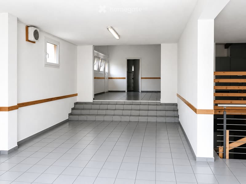 Empty hallway with tiled floor, white walls, and a staircase leading to another level. Features include a small window, an air conditioning unit, and a railing on the staircase.
