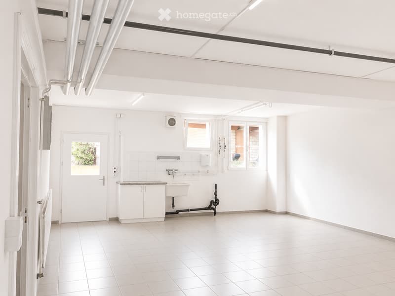 Completely empty room with white tiled floor and walls, a sink, and a door leading outside.