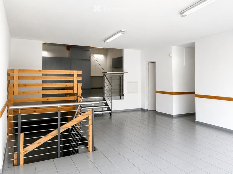 Modern hallway with white walls, grey tiled floor, and a staircase with wooden and metal railings leading to another level. The area is well-lit with recessed lighting and features a simple, clean design with a door visible on the right side.