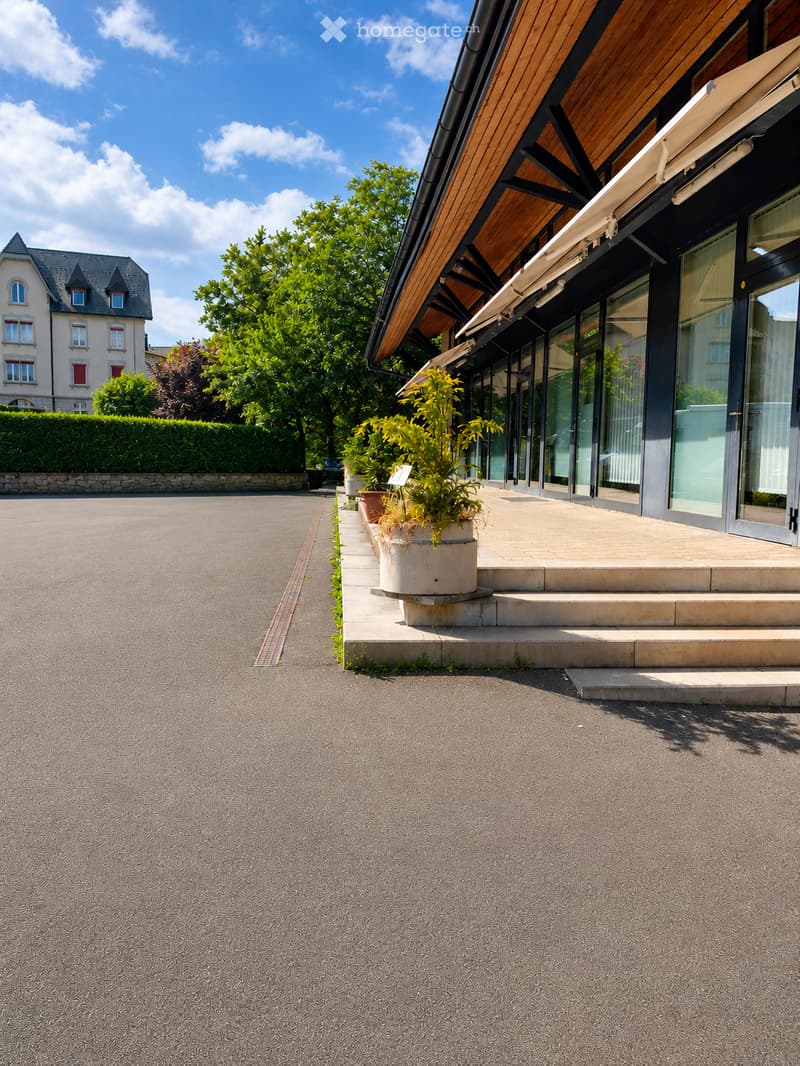 Modern building entrance with glass doors, steps, potted plants, and a paved driveway. A traditional house is visible in the background.