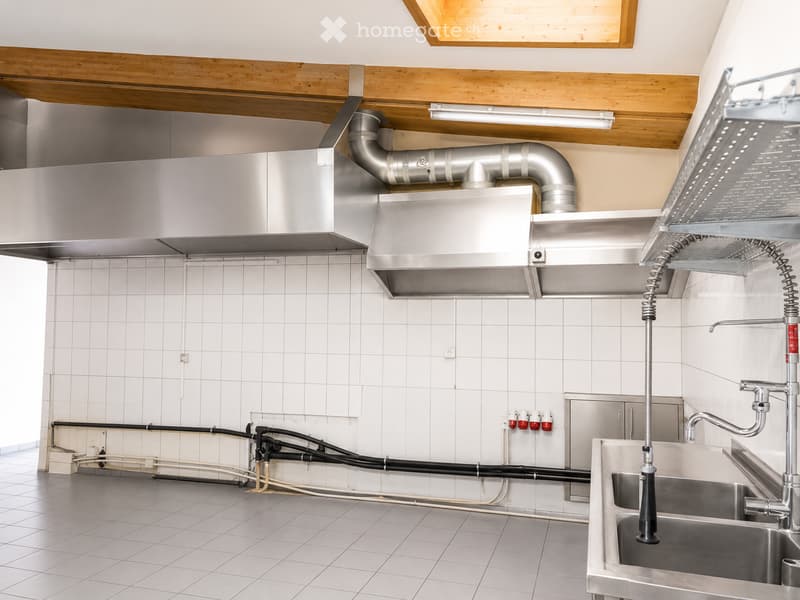 Commercial kitchen with stainless steel sink, faucet, range hood, and exposed pipes and wiring.