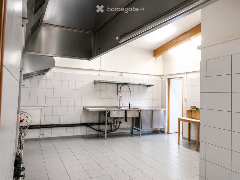 Commercial kitchen with white tiled walls and floor, stainless steel sink, faucet, shelves, and a small wooden table with boxes.