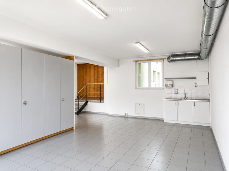 Empty room with white tiled floor, white walls, and a small kitchenette area with sink and cabinets. A staircase leads to another level.