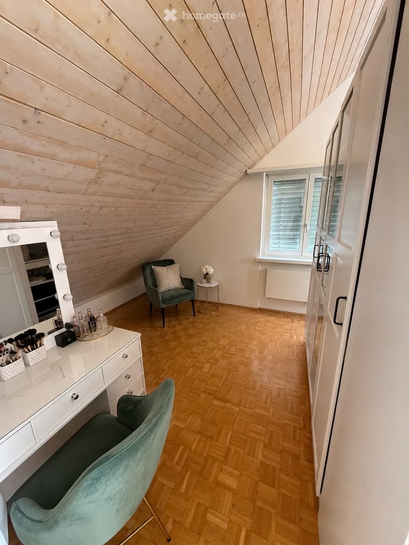 A cozy, well-lit room with wooden flooring and slanted wooden ceiling. Features include a vanity with a mirror, a green armchair, a small round table with a vase, and a large window with blinds.