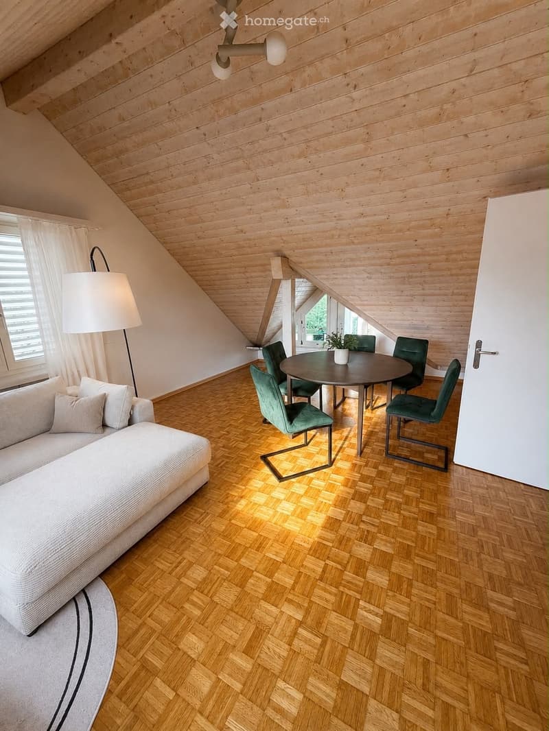 Modern attic room with wooden slanted ceilings, herringbone wooden flooring, a white sofa, a round dining table with green chairs, a floor lamp, and a potted plant on the table.