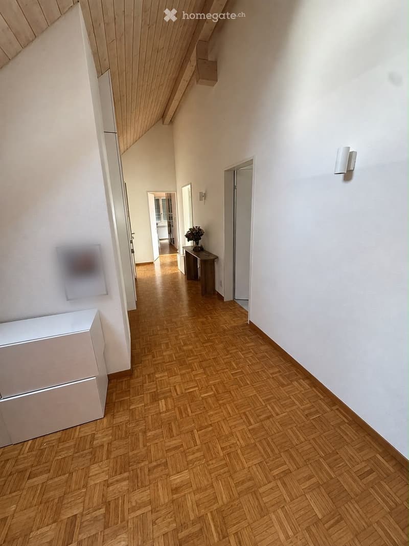 A narrow hallway with wooden herringbone flooring, white walls, and several doors leading to other rooms. A small table with a vase of flowers is visible at the end of the hallway.