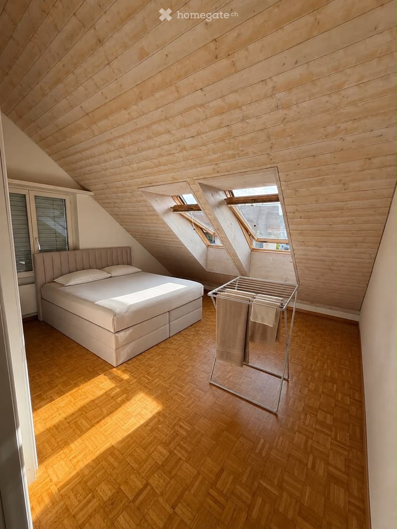 Cozy attic bedroom with wooden slat ceiling, herringbone wooden floor, large skylights, and a neatly made bed with pillows and a blanket.