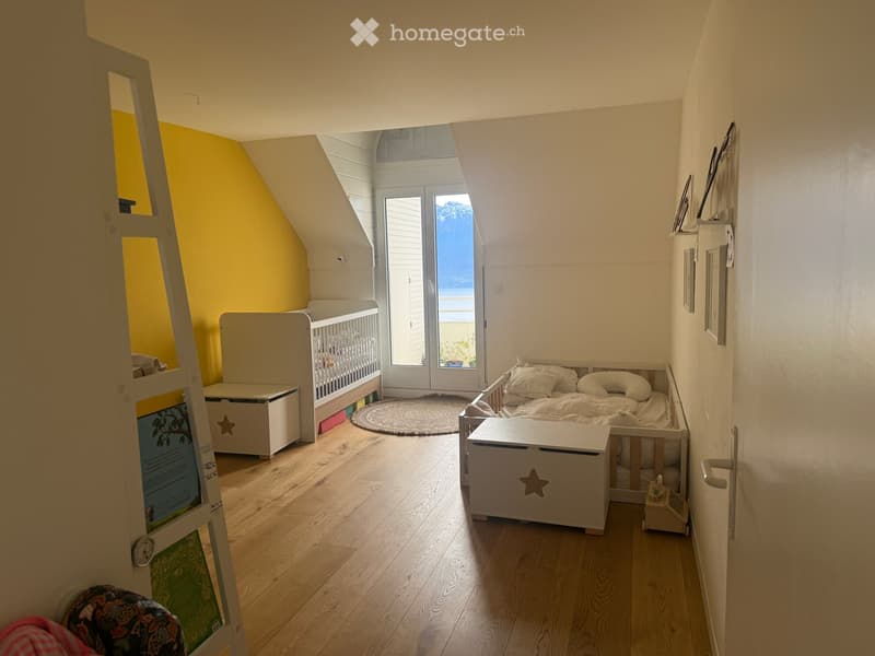 Cozy children's bedroom with a crib, daybed, toy boxes, and a round rug, featuring wooden flooring and large windows with a view.