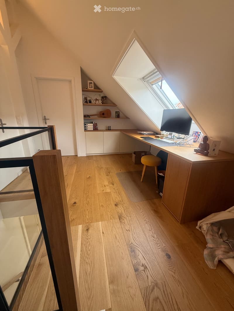A cozy attic room with wooden flooring, a large window, a desk with a laptop, a stool, shelves with books and a guitar, and a small bed tucked into the corner.