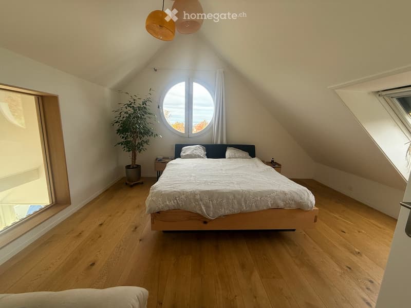 Cozy attic bedroom with wooden floors, slanted ceilings, large round window, and a bed with white bedding.