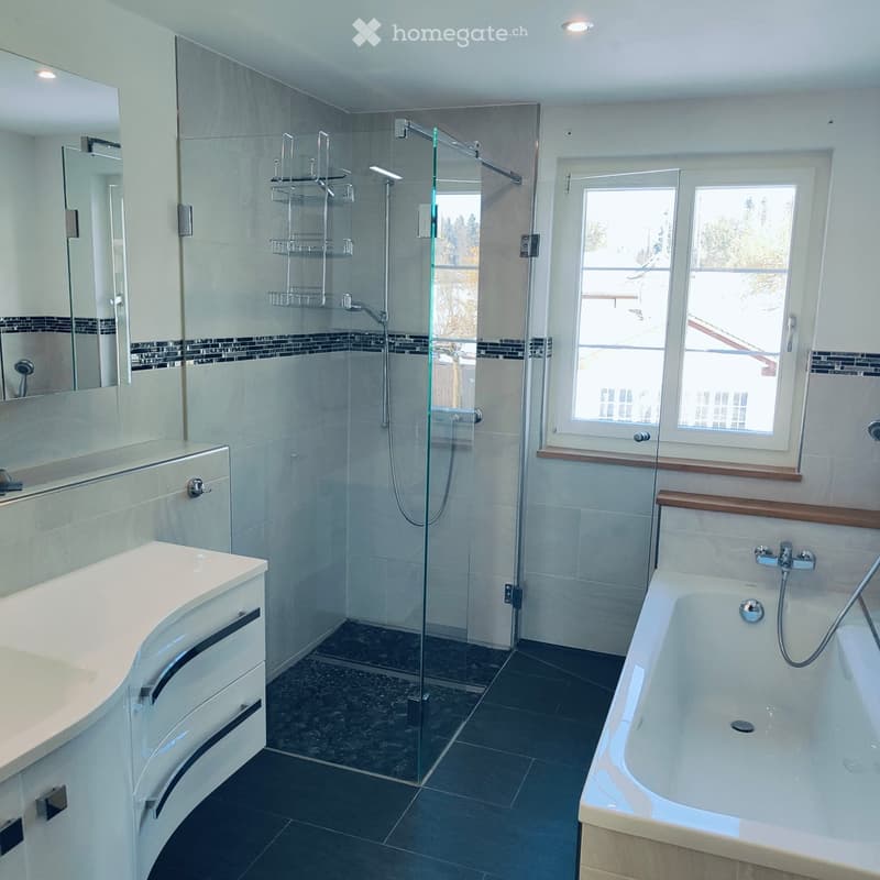 Modern bathroom with a bathtub, sink, mirror, and glass-enclosed shower. Features dark tiled floors and light-colored walls.