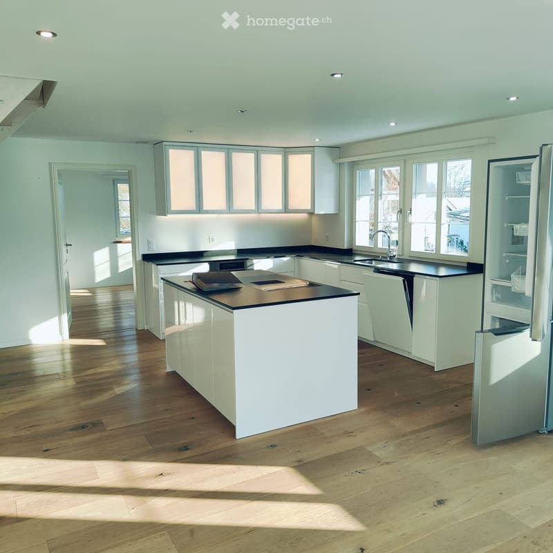 Modern kitchen with white cabinets, black countertops, wooden flooring, and large windows allowing natural light.