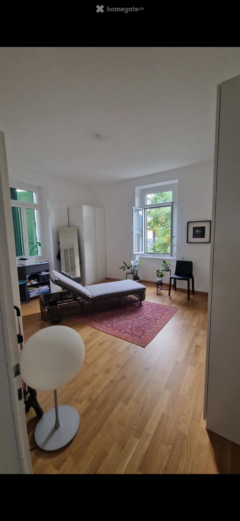 Bright room with wooden flooring, large windows, daybed, chair, stool, mirror, potted plants, cabinet, and picture frame.