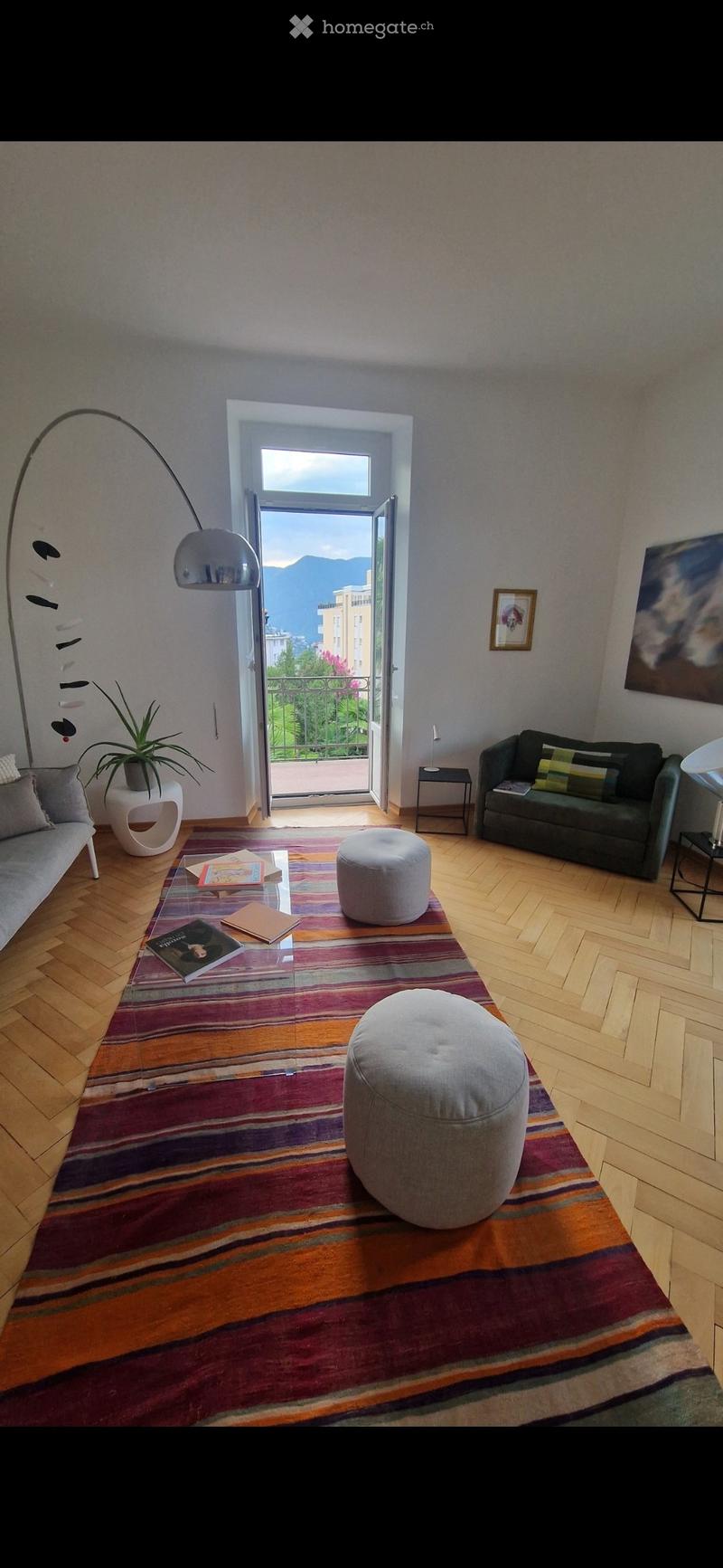 Modern living room with wooden flooring, large window opening to a balcony with a scenic mountain view, sofa, ottomans, coffee table, floor lamp, side table, plant, and artwork.